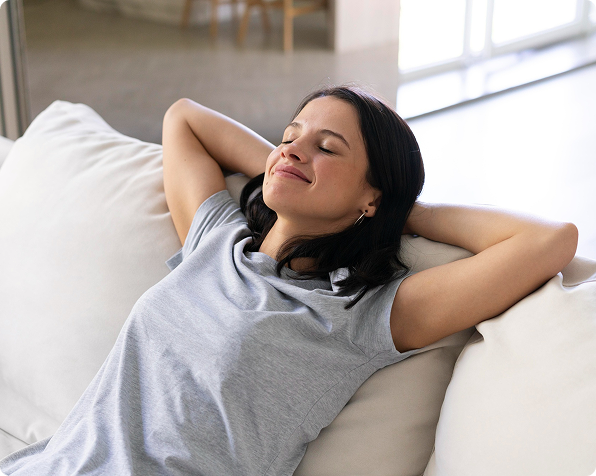 A woman in a gray t-shirt relaxes on a white couch with her hands behind her head and eyes closed, appearing content after organizing her space with self storage.
